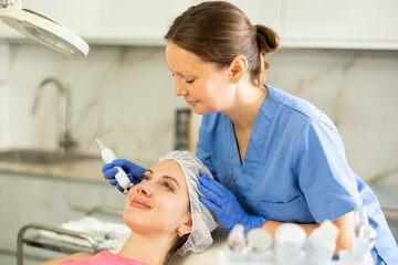 Adult woman cosmetologist performs hardware facial microcurrent procedure to young female patient