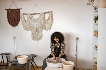 Young female potter creating ceramic dishes on pottery wheel in cozy studio. She is focused and smiling, enjoying the creative process.