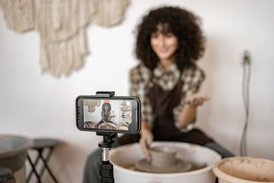 Young woman demonstrating pottery techniques on potter's wheel while recording vlog. Focus on smartphone capturing video in foreground.