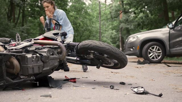 Collision between motorcycle and car on the road. Girl driver hit motorcyclist and uses her phone to call for help. Broken and wrecked motorcycle on road