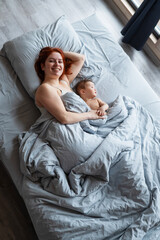 Top view of a red-haired Caucasian woman lying in bed with her baby son. Vertical photo.