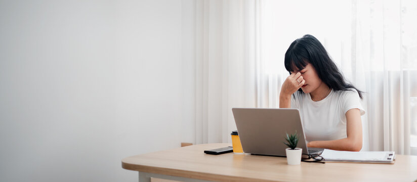 Young woman feeling stressed while working from home at a desk with a laptop and coffee in a modern minimalist home office setting.