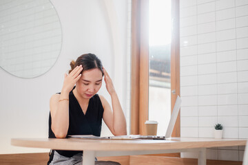 Asian woman experiencing stress while working from home, sitting at a minimalist desk with a laptop...