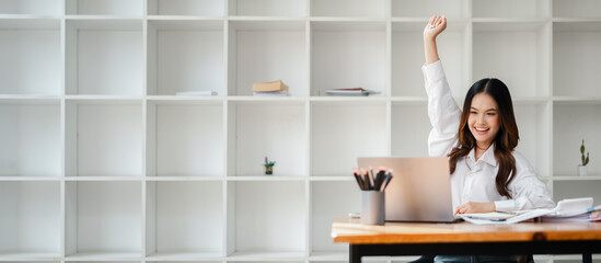 Happy young woman raising her hand in celebration while working on a laptop in a modern office with...