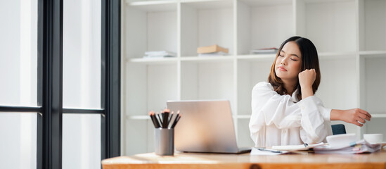 A young woman takes a break from work, stretching her arms at a home office desk with a laptop and...