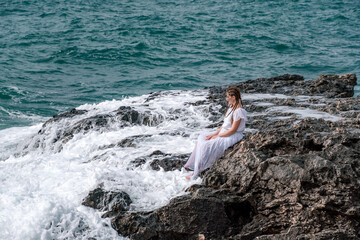 Fototapeta premium A woman in a storm sits on a stone in the sea. Dressed in a white long dress, waves crash against the rocks and white spray rises.