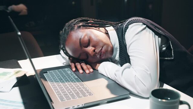 Tired and asleep, black woman with laptop in office at night for burnout, fatigue and overtime. Exhausted, female person or workaholic and sleeping or dreaming at desk with computer for deadline