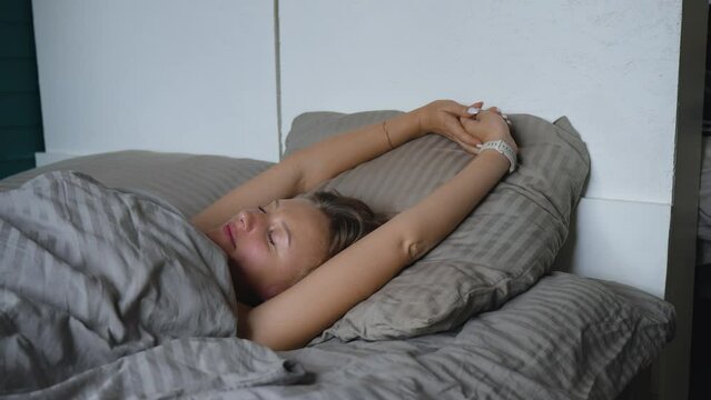 A morning moment of rest and relaxation: a young blonde woman lying in bed, turning over gently stretching and waking up.