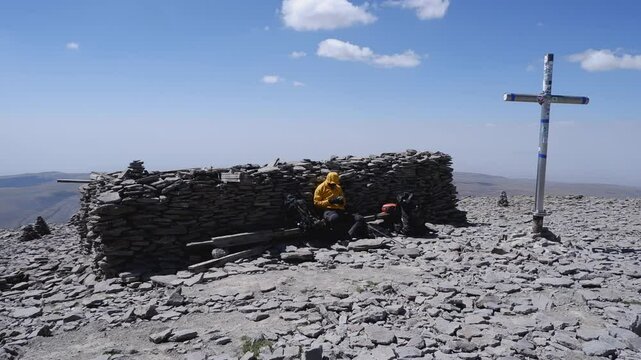 Climber rests by huge stone cairn near crucifix on mountain summit