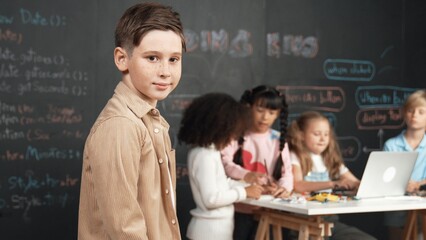 Smart boy smiling at camera while diverse friend working or learning engineering code or prompt in...