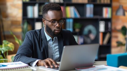 Obraz premium Concentrated African businessman in glasses using laptop in a modern library setting