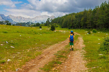 Obraz premium hiking in the mountains. A little boy with a backpack walks along a path against a background of mountains and clouds. Active healthy lifestyle on weekend hike journey.