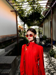 Stylish woman in red dress and sunglasses striking a pose in a sunny courtyard