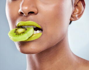 Cosmetics, woman and kiwi fruit in mouth for wellness, beauty and healthy eating in grey studio background. Detox, organic food and female person green lips isolated against backdrop for dermatology