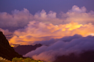 Under the veil of night, the dramatic clouds above Pico do Arieiro dance in a mesmerizing display, illuminated by the vibrant hues of magenta and orange city lights below.