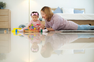 Cute asian mother take care and play with happy little baby with many wooden toy on the floor of bedroom home.