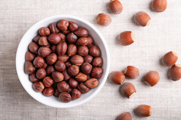 Hazelnuts in a white bowl on linen fabric. Whole, dried and shelled nuts of Corylus avellana, on the right side in the shells. Ready to eat as snack, and also used for baking. Close-up, from above.