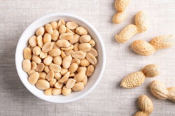 Roasted and salted peanuts in a white bowl on linen fabric. Ready-to-eat snack food, made from fruits of Arachis hypogaea, also known as groundnut, goober, pindar or monkey nut. Close-up from above.