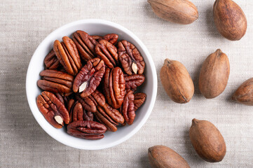 Pecan nuts in a white bowl on linen fabric. Dried pecan halves and whole pecans in shells, seeds and nuts of Carya illinoinensis. Ready to eat as snack, and also used for baking. Close-up, from above.