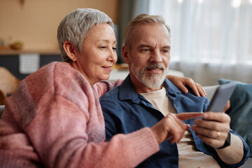 Portrait of smiling senior couple using phone together and tapping screen with mobile app at home