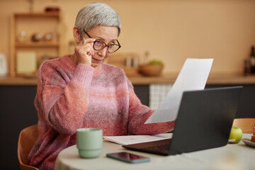 Portrait of Asian senior woman wearing eyeglasses reading document and doing taxes at home copy...