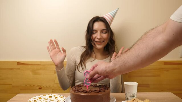 A girl sits in front of a table with a festive cake, into which a candle in the form of the number 26 is stuck. The concept of a birthday celebration. Slow motion