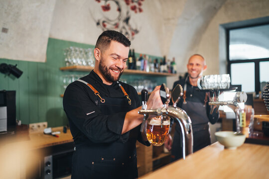 Stylish bearded barman dressed black uniform smiling at camera, beer tapping at bar counter and waiter with tray. Successful people teamwork, friendship, brewing and restaurant industry concept image