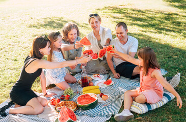 Big caucasian family on picnic blanket on the in city park green grass. They rose-up red juicy watermelon pieces and clinking them like glasses. Family values and outdoor activities concept.
