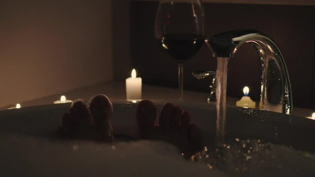 Toes sticking out of water of a bubble bath at night with candles and red wine