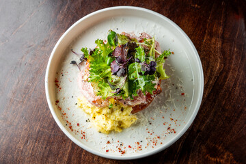 A gourmet salad with mixed greens, grated cheese, and dressing on a sliced tomato, presented in a white bowl.