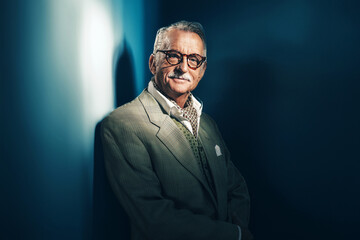 Obraz premium Senior man with mustache and glasses in suit with choker standing by a wall at night.