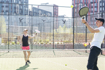 Portrait of positive young woman and adult man standing on padel tennis court, holding racket and ball, smiling 