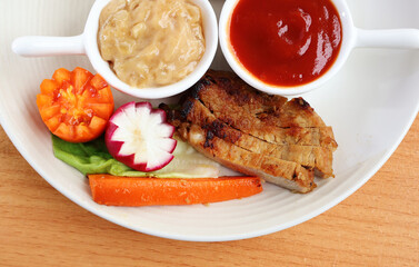 Roast pork, carrots, radish, tomatoes and a bowl of sauce in a white porcelain plate on a wooden table.

