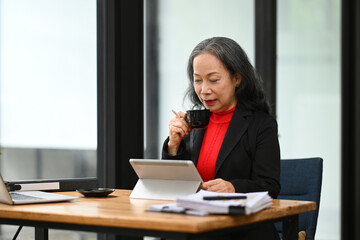Professional senior businesswoman drinking coffee and using digital tablet at office desk