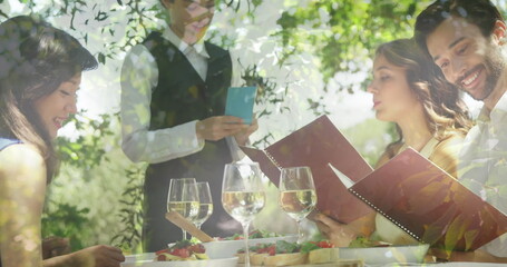 Image of foliage over caucasian friends and waitress ordering wine at restaurant