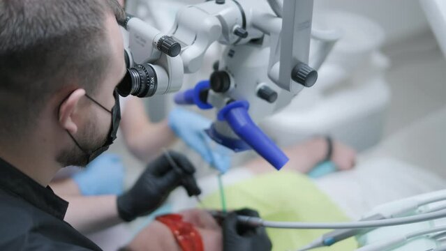 Male dentist working with dental microscope in modern stomatology clinic. Doctor using microscope for root canal treatment. Stomatologist performs a surgical operation. Cosmetic dentistry and surgery.