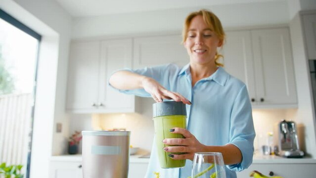 Woman pouring and drinking fresh healthy green smoothie made from apples,banana and celery from blender into glass in kitchen at home - shot in slow motion