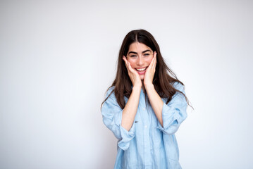 Young woman with long dark hair is smiling and touching her face with both hands on a white background