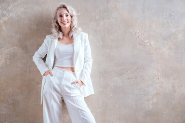 Portrait of young beautiful blond woman wearing nice trendy white suit. Fashion model posing in studio. Fashionable female posing near grey wall. Cheerful and happy