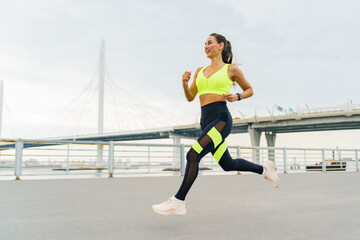A woman jogs energetically along an urban waterfront, smiling brightly in vibrant sportswear under a clear sky.