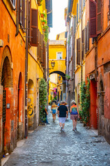 Tourists walk through a narrow, cobblestone alley in Rome, Italy. The buildings are brightly colored and adorned with plants and flowers. The alley leads to an archway, providing a glimpse of the city