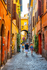 Naklejka na ściany i meble Tourists walk through a narrow, cobblestone alley in Rome, Italy. The buildings are brightly colored and adorned with plants and flowers. The alley leads to an archway, providing a glimpse of the city
