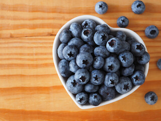 Organic ripe fresh blueberries in heart shape white bowl on wooden background. Concept for healthy eating and nutrition, Top view with space for your text. Superfood..