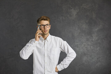 Young handsome serious man talking on mobile phone with confident expression on smart face. Caucasian young man in white shirt and glasses stands on gray background. Phone talk concept. Banner.