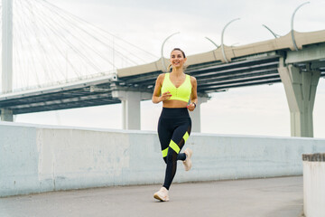 A woman jogs along a waterfront path, smiling confidently in vibrant sportswear, with a modern bridge in the background.