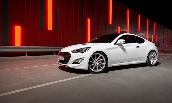 white Hyundai Genesis Coupe parked near illuminated red wall at night. Front three quarter view of sporty korean coupe in urban setting. Kyiv, Ukraine - June 07 2015.