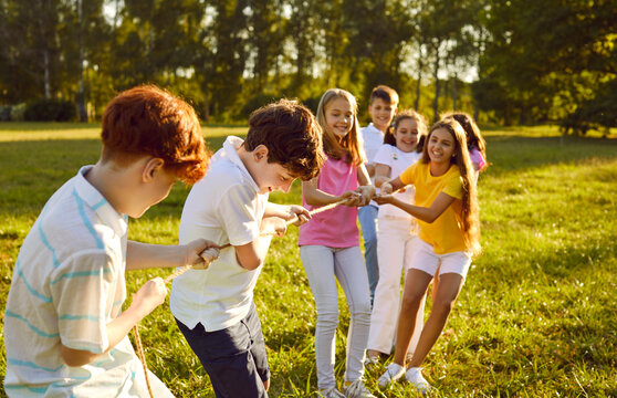 Group of a happy joyful smiling children playing together in tug-of-war with a rope in the park on summer holidays. Cheerful school kids having fun on green grass outdoors in the camp.