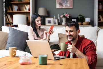 Young happy newlywed couple sitting at home, searching the internet on laptop and smartphone, search travel destination for travel. Man and woman online booking hotel for vacation.