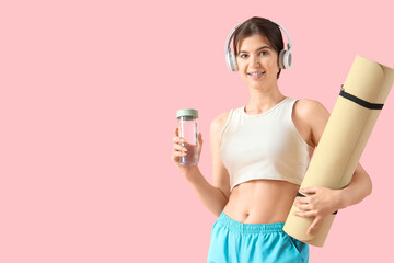 Sporty young woman in headphones with fitness mat and water bottle on pink background