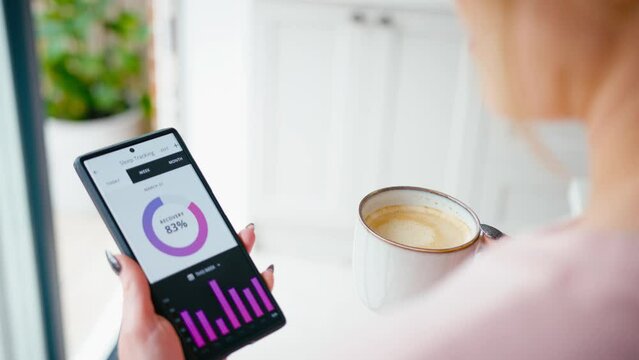 Close up of woman drinking coffee looking at sleep tracking app on mobile phone at home - shot in slow motion
